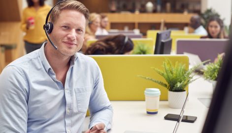 Portrait Of Businessman Wearing Headset Talking To Caller In Busy Customer Services Centre